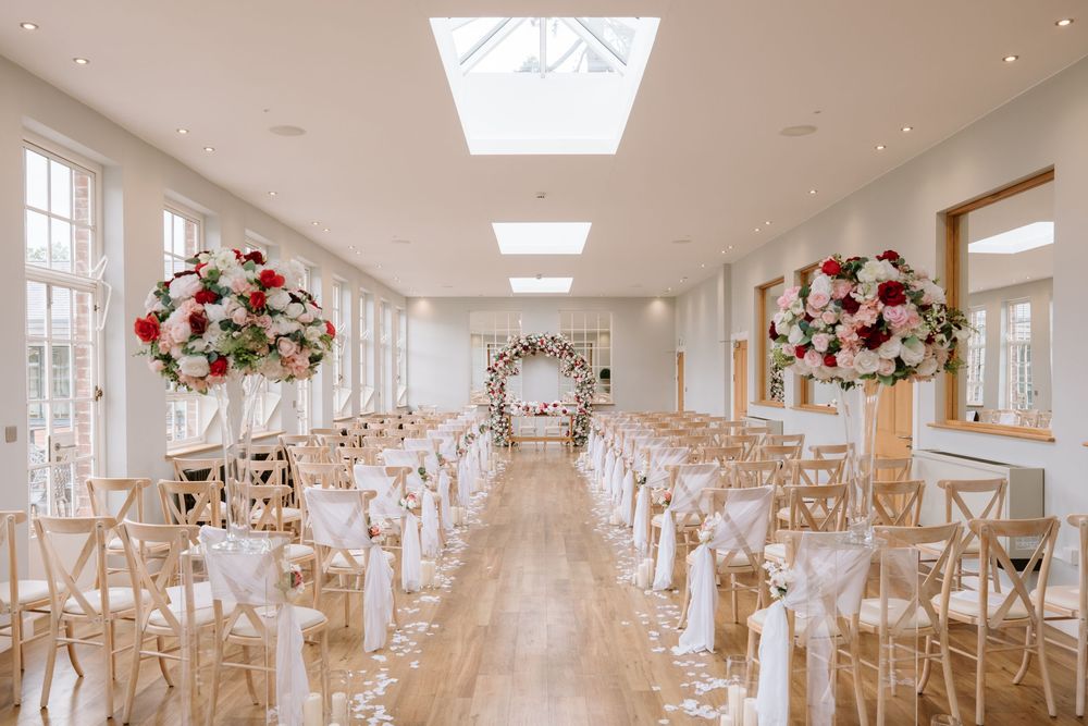A bright white wedding reception hall decorated with wooden chairs, floral arrangements, and natural light from skylights.