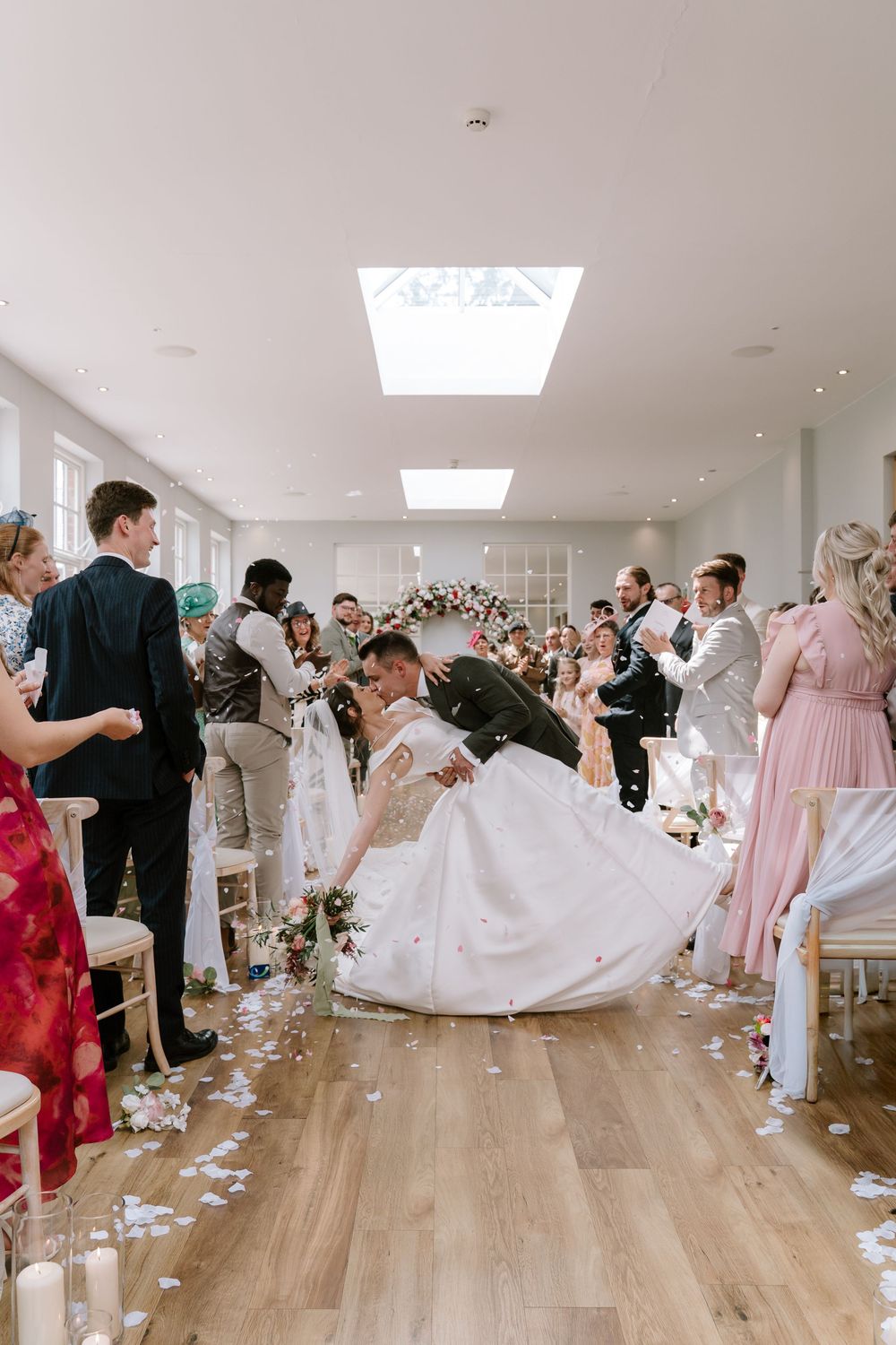 A romantic wedding ceremony moment in a bright indoor venue with skylights and wooden floors surrounded by guests.