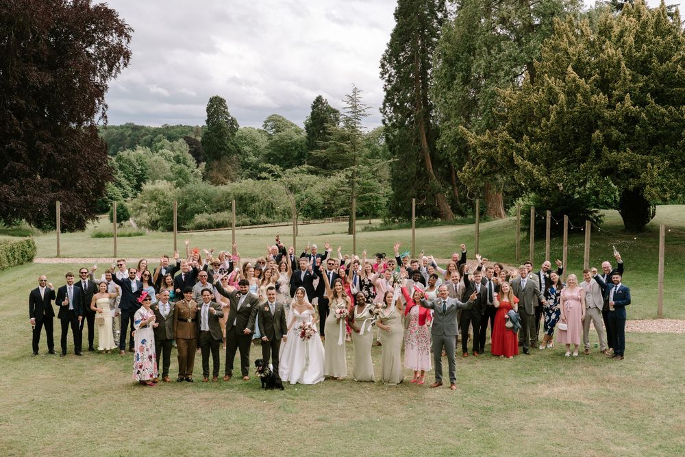 Large Cotswold wedding party gathered on a green lawn surrounded by trees for a group photo on an overcast day.