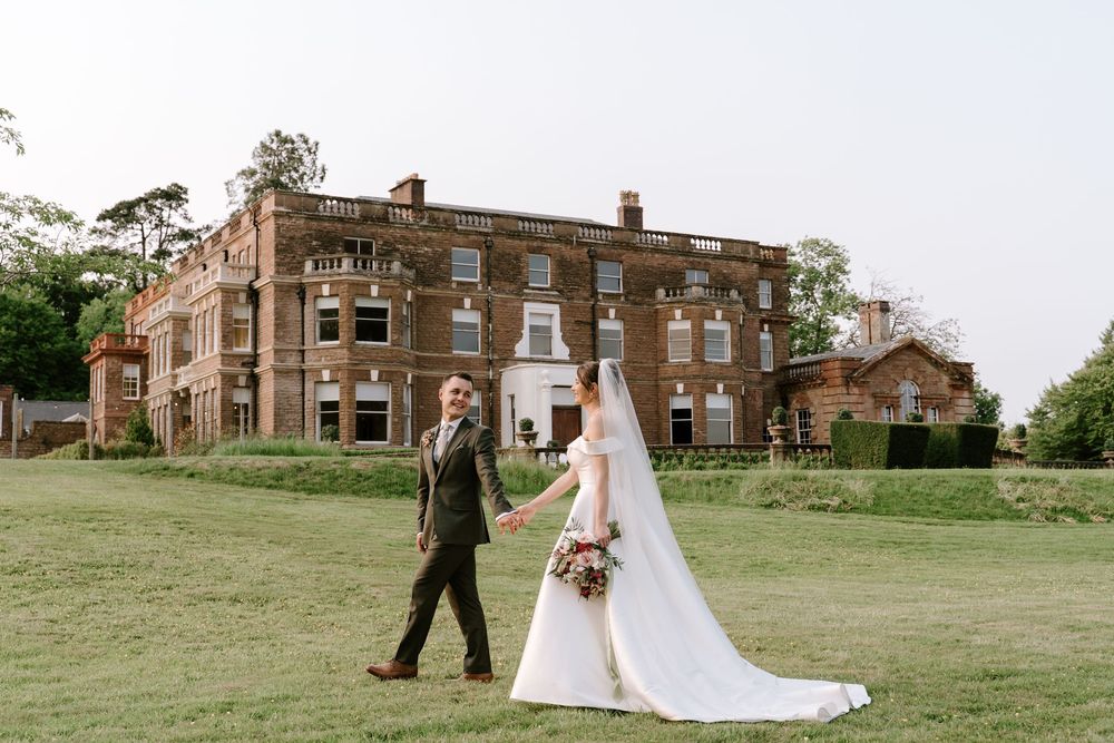 A Worcestershire wedding couple walks together on a lush green lawn with Bredenbury Court Barns in the background.