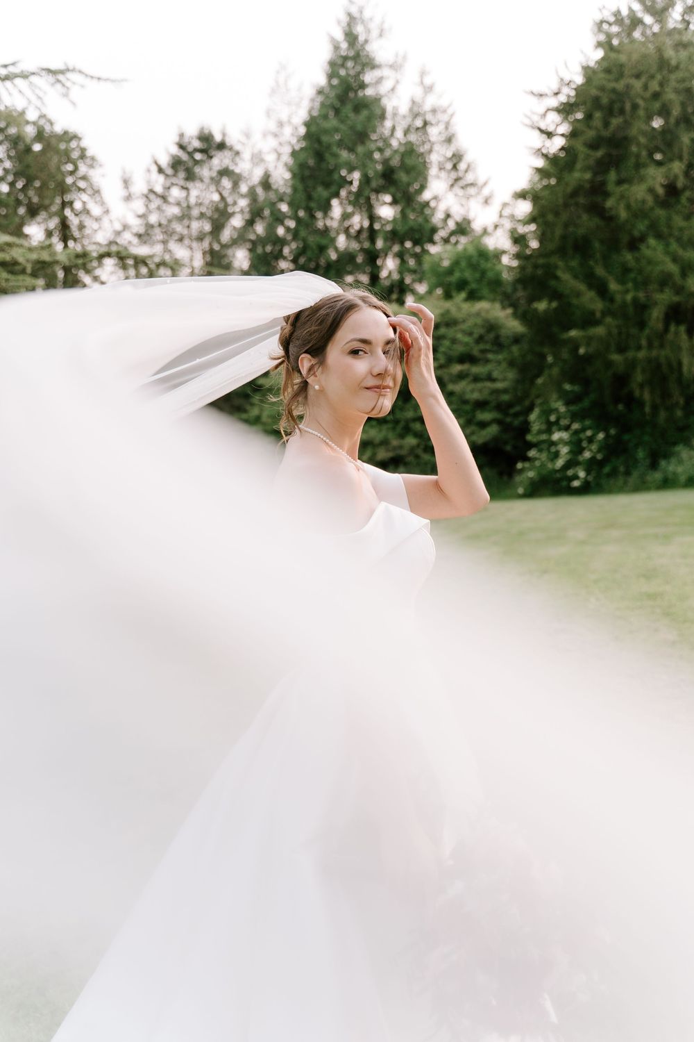 A flowing white wedding veil billows in the breeze against a backdrop of evergreen trees on a sunny day.