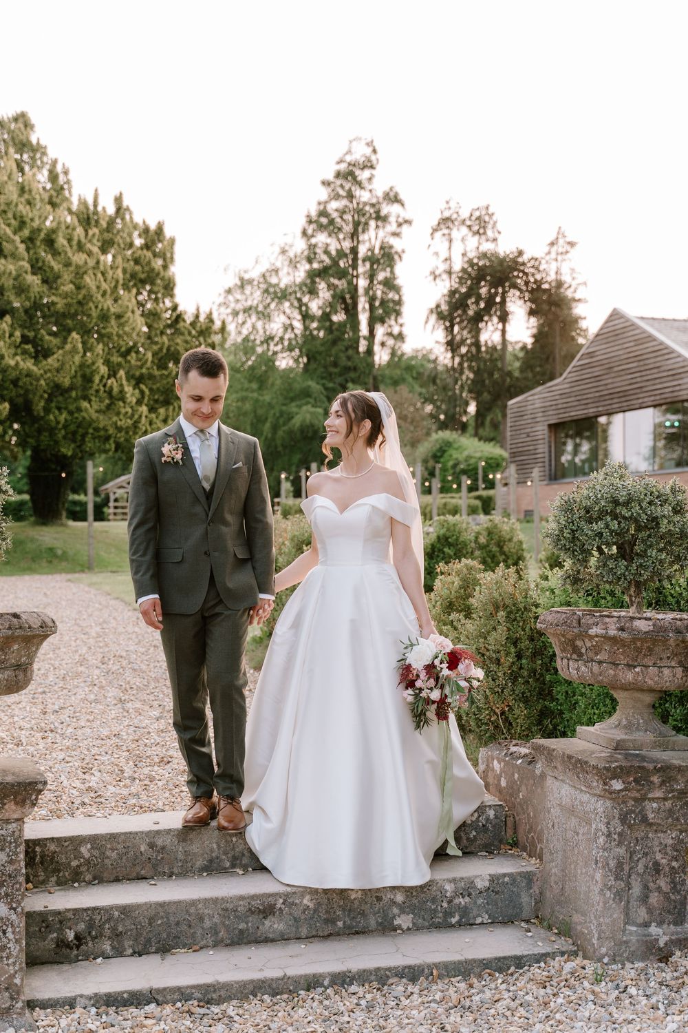 A Worcestershire couple in wedding attire stands on stone steps in a garden setting with gravel paths and greenery.