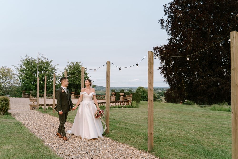 A couple stands on a gravel path under string lights at  a Worcestershire venue with lush green grounds.
