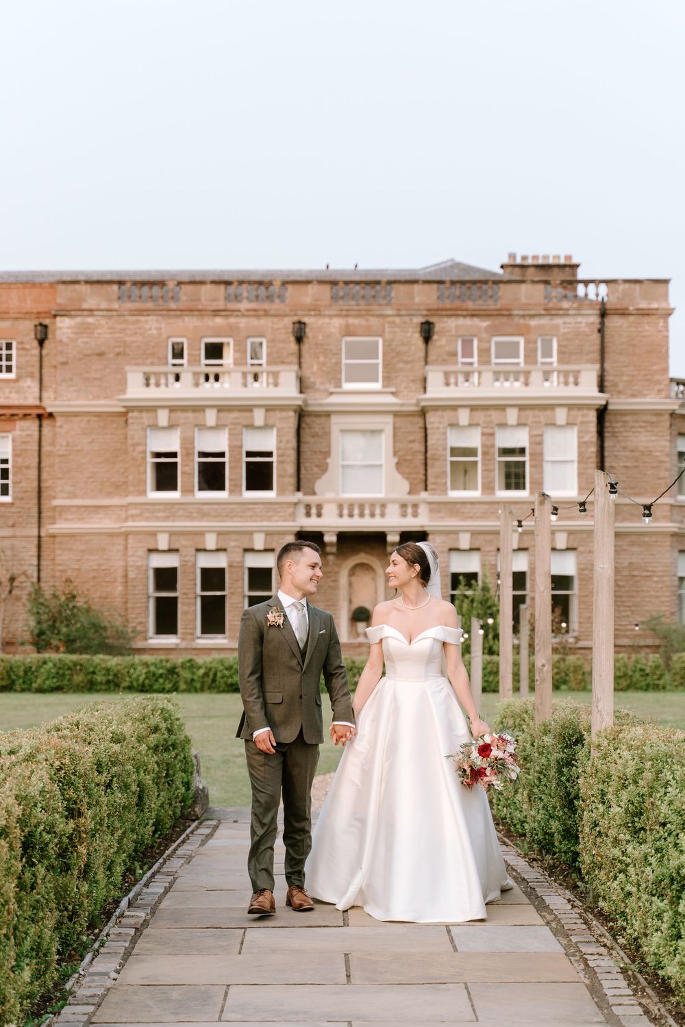 A Cotswold wedding couple walks together on a stone path outside a historic brick building on their wedding day.