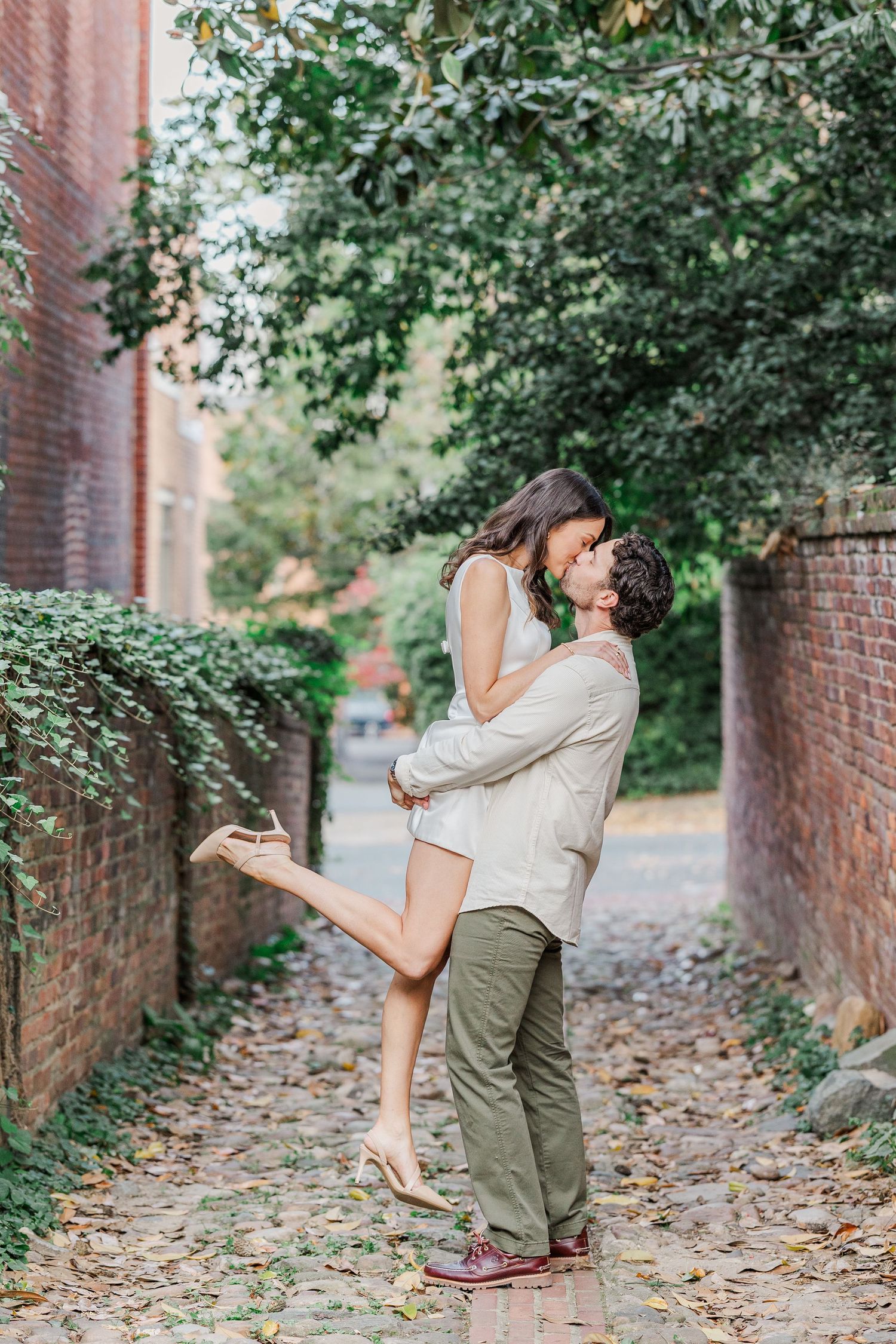 A couple embraces and spins joyfully in a brick-lined alley with autumn leaves scattered on the ground.
