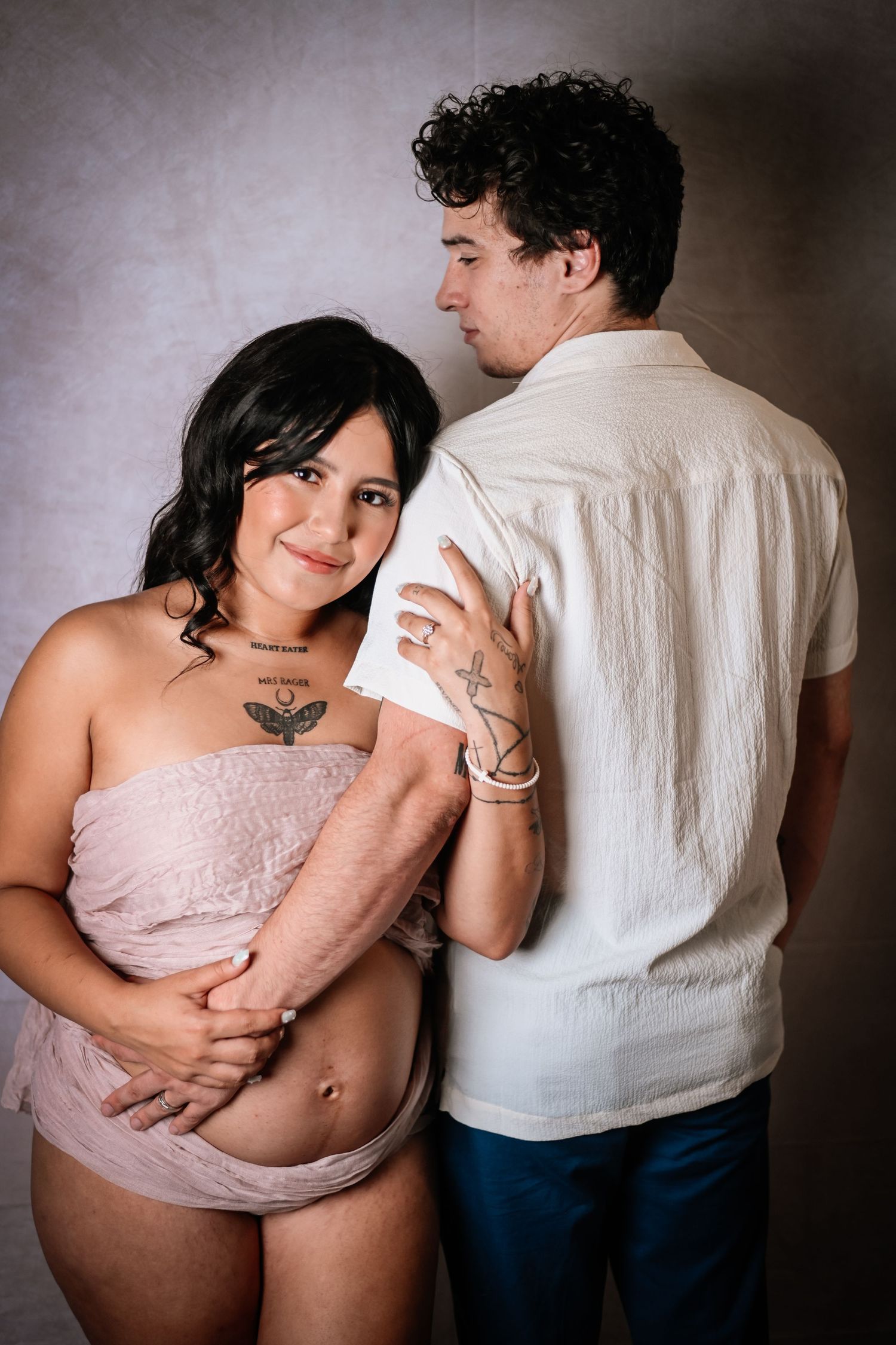 Maternity photo series showing couple embracing in intimate poses against white wall in soft lighting.