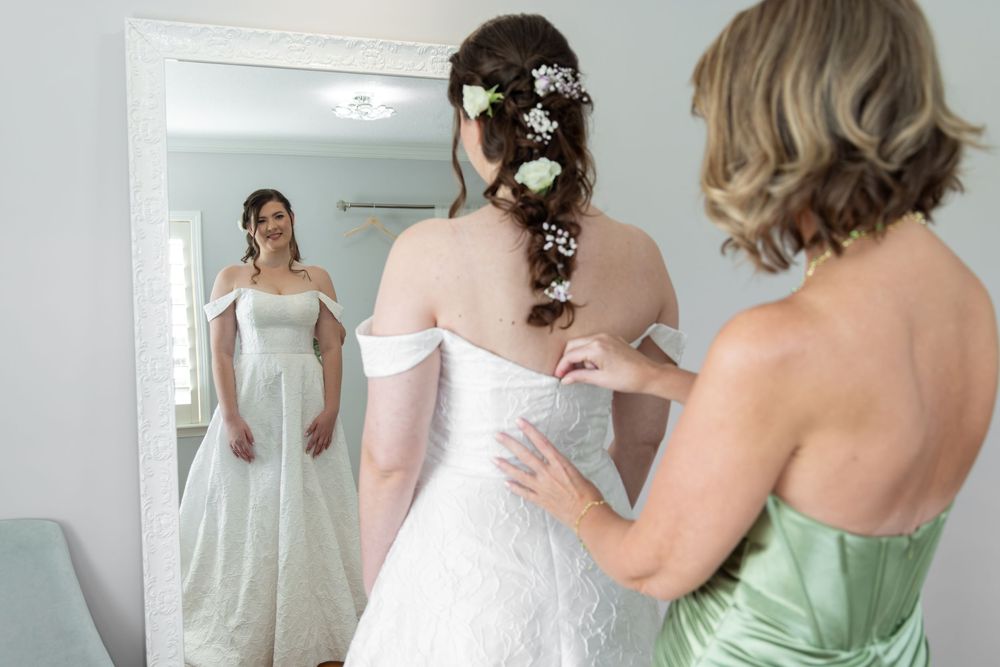 Bridesmaids in pastel green dresses help adjust a bride's white wedding gown in a sunlit dressing room.