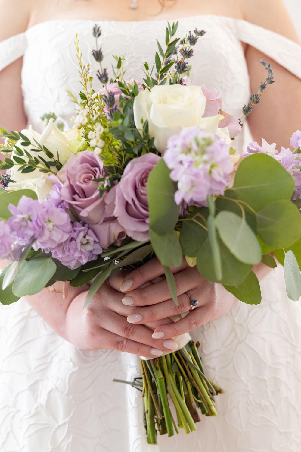 Close-up view of a bridal bouquet featuring purple roses, white flowers and eucalyptus leaves against a white dress.