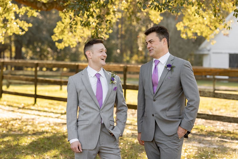 Two people in gray suits and pink ties laugh together while standing outdoors in autumn sunlight.