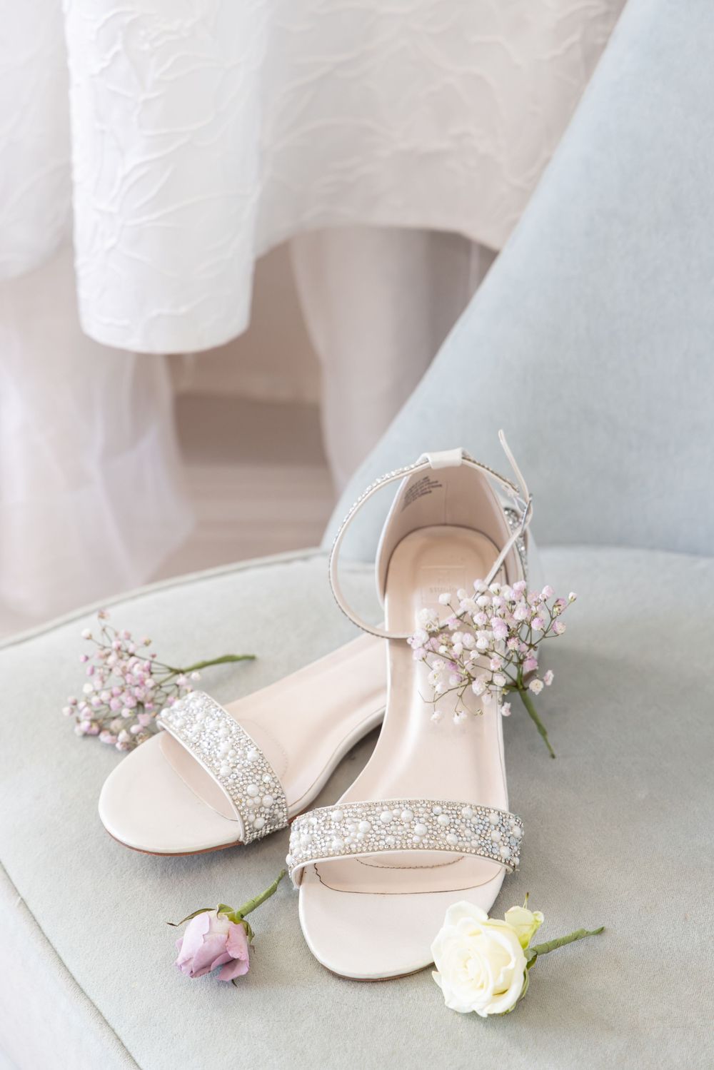 Elegant white bridal shoes with delicate floral details and crystals displayed with small flowers against a light backdrop.