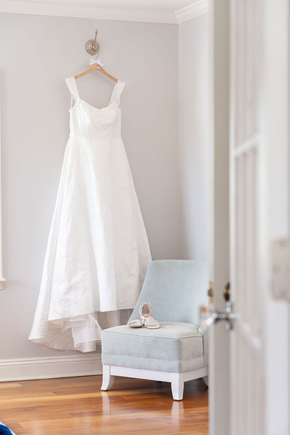 An elegant white wedding dress hangs on a hanger next to a light blue ottoman chair in a bright room.