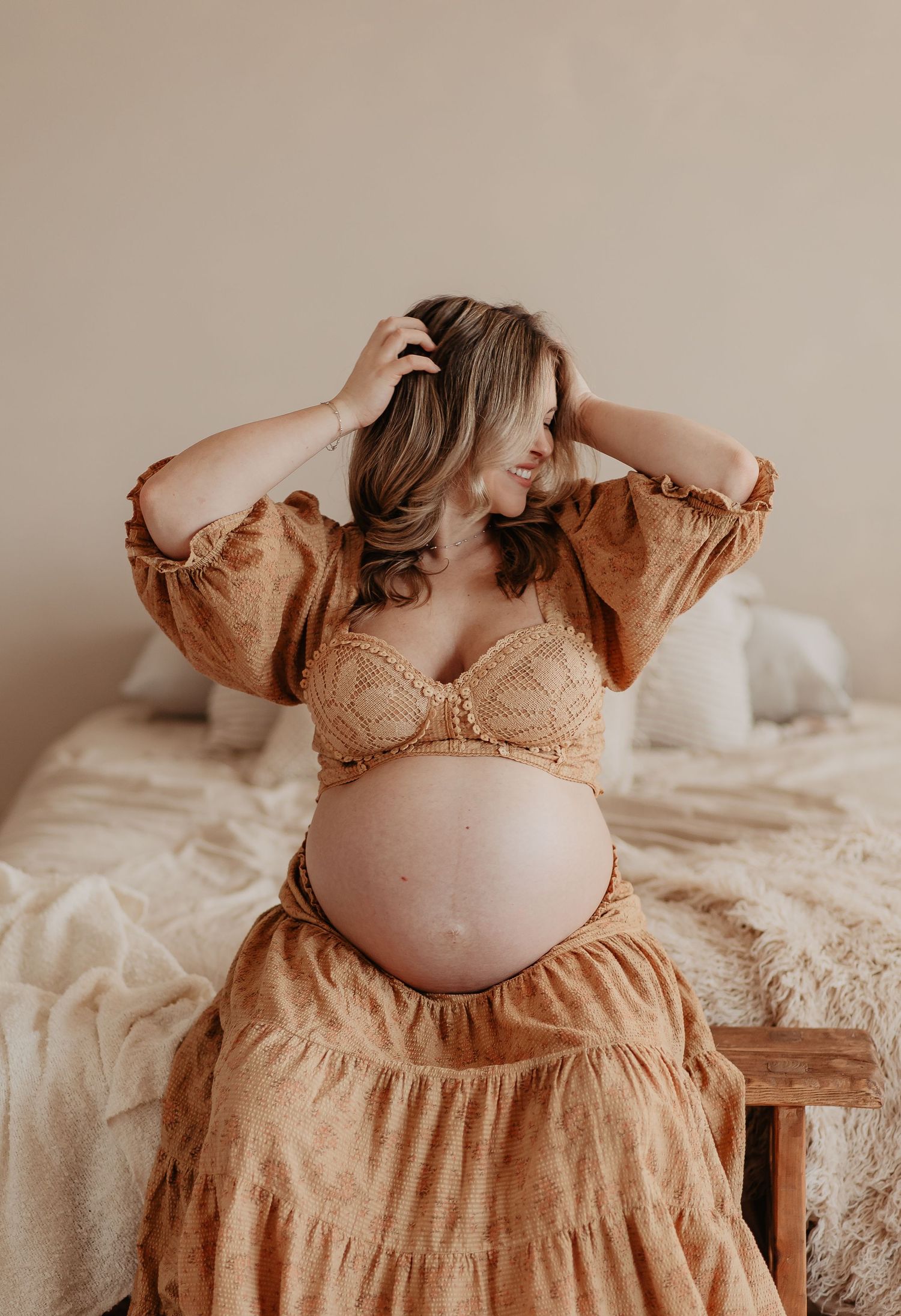 A pregnant woman in a white sweater relaxes on beige bedding while showing her bare belly in a maternity photoshoot.