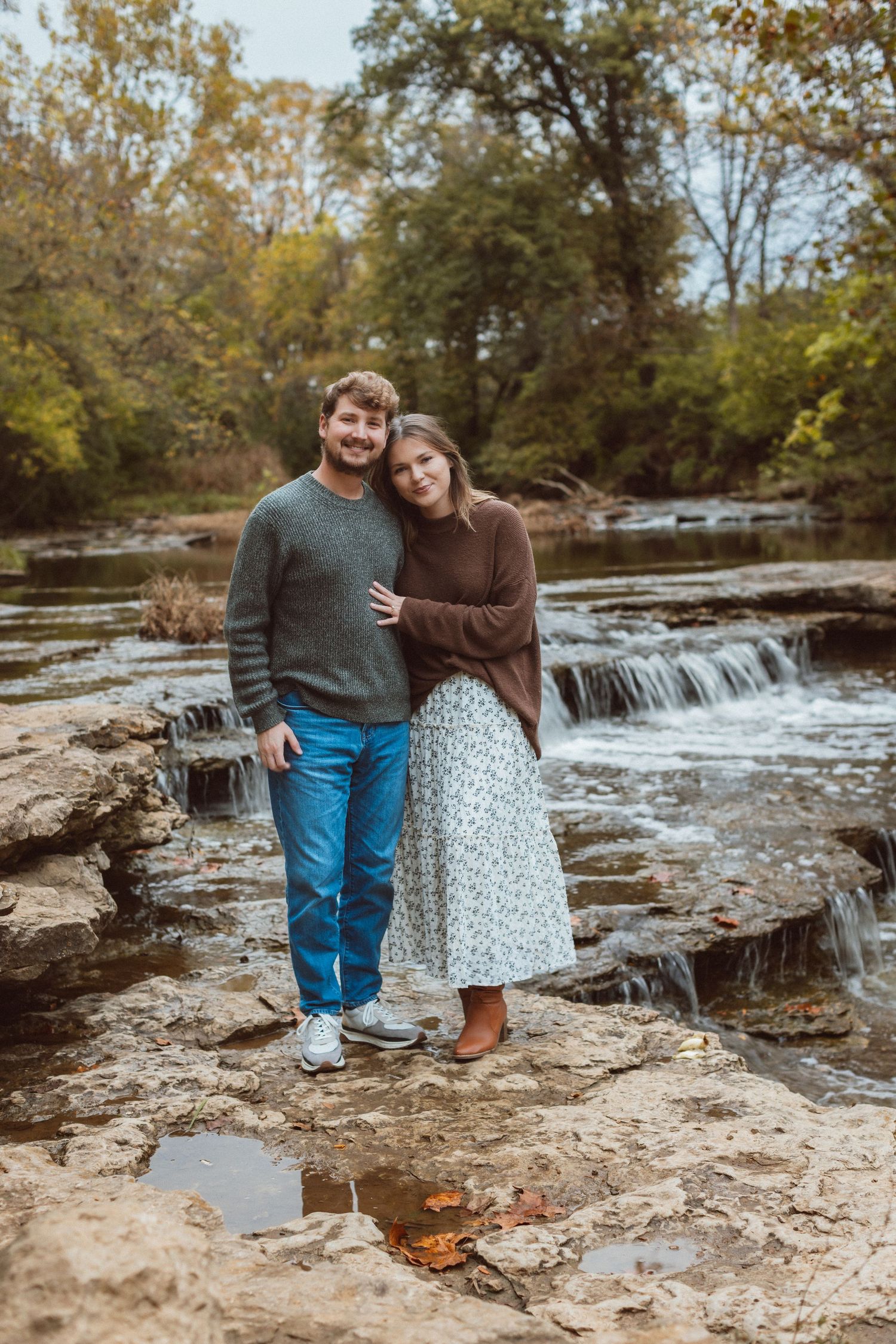 A man and woman stand close and smile for the camera in front of a waterfall during their Kansas City engagement photo session.