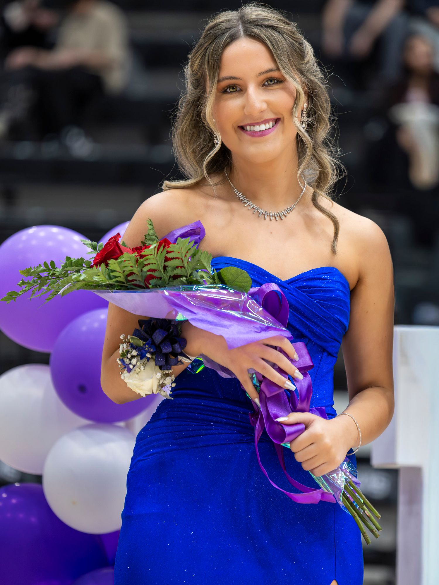 Woman in royal blue strapless gown smiles while holding purple-wrapped flowers and standing near purple and white balloons.