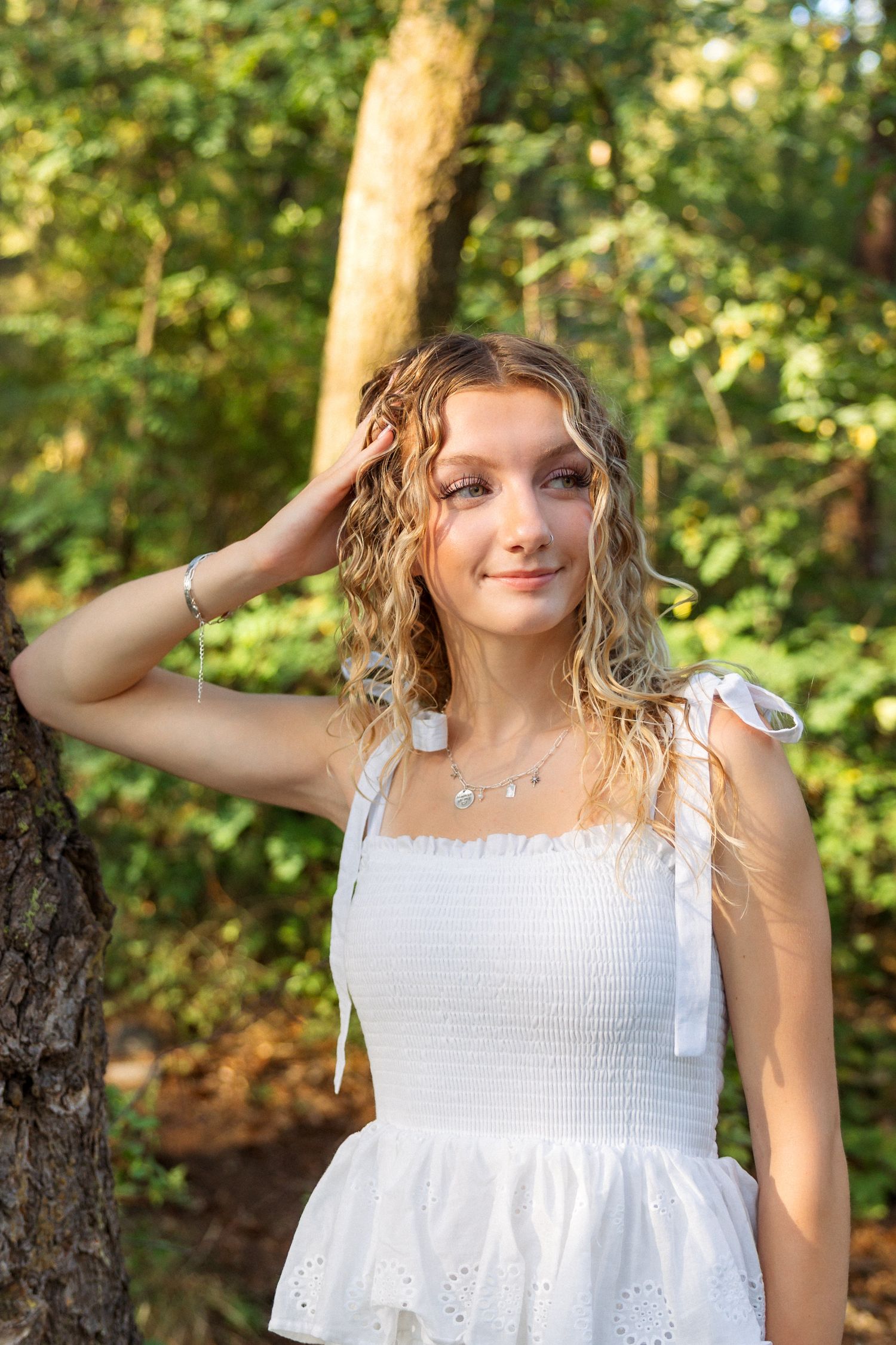 A person in a white summer dress poses against a sunlit forest backdrop with green foliage.