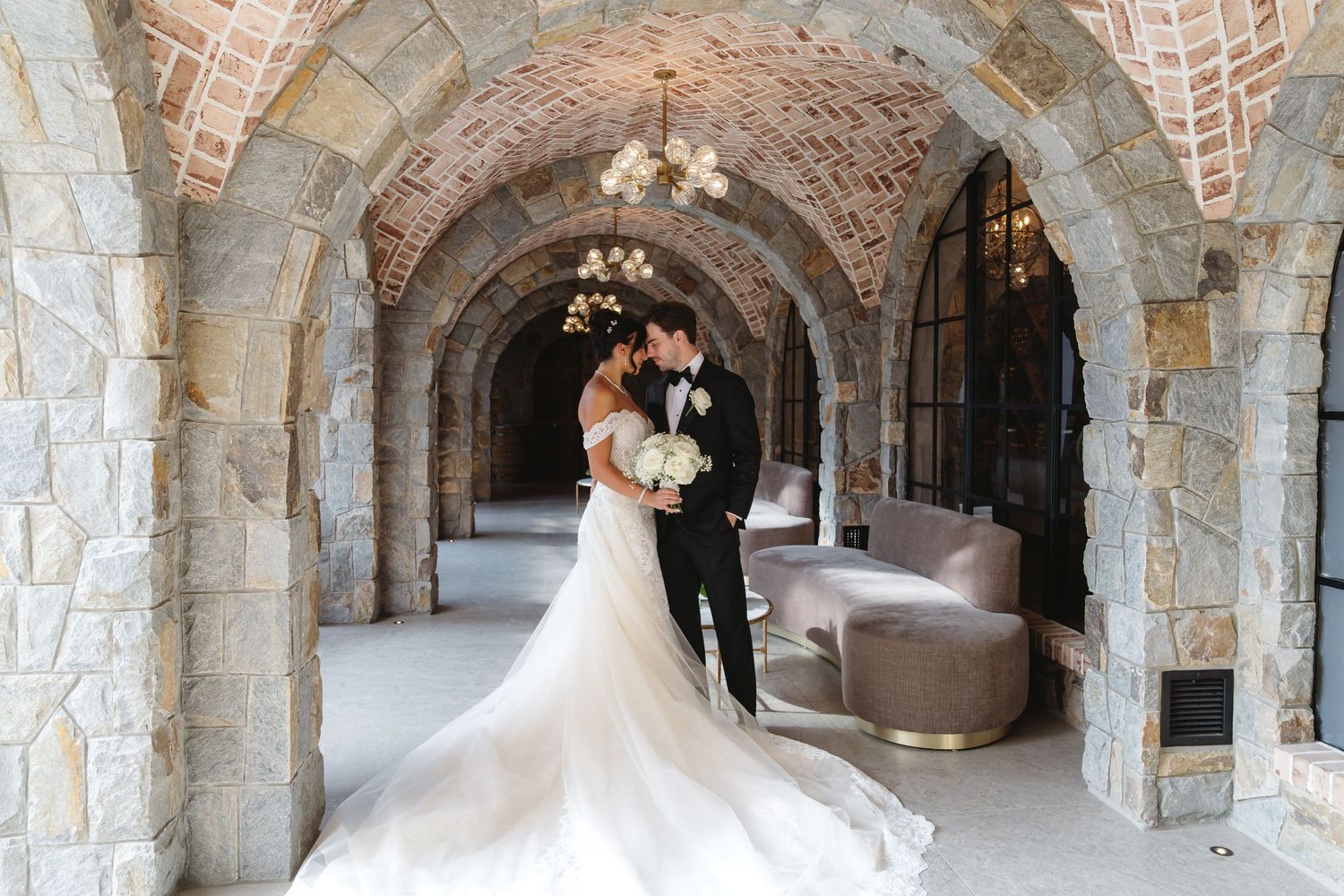 A romantic wedding moment captured under a stone archway corridor with a flowing white dress trailing behind.