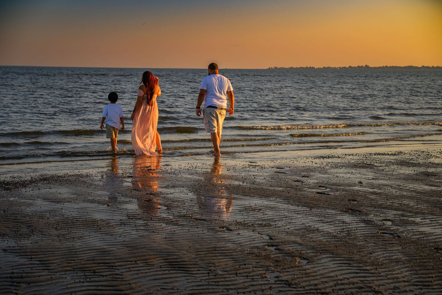 A family of three walks along a reflective shoreline at sunset, with warm golden light over the water.