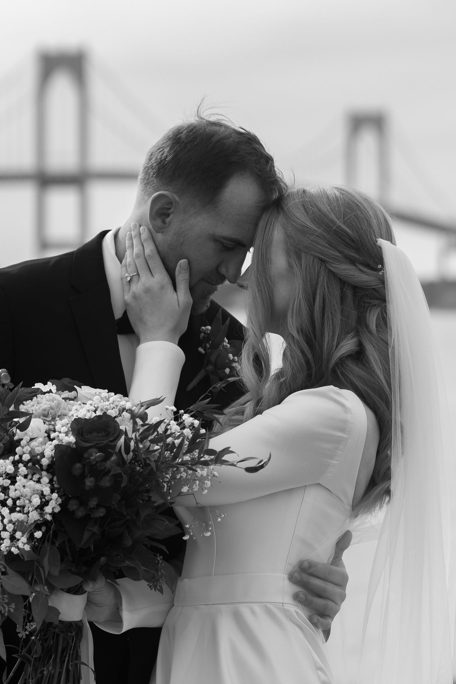 A romantic black and white wedding photo showing a tender moment between newlyweds near the Newport bridge in Rhode Island with a bouquet.