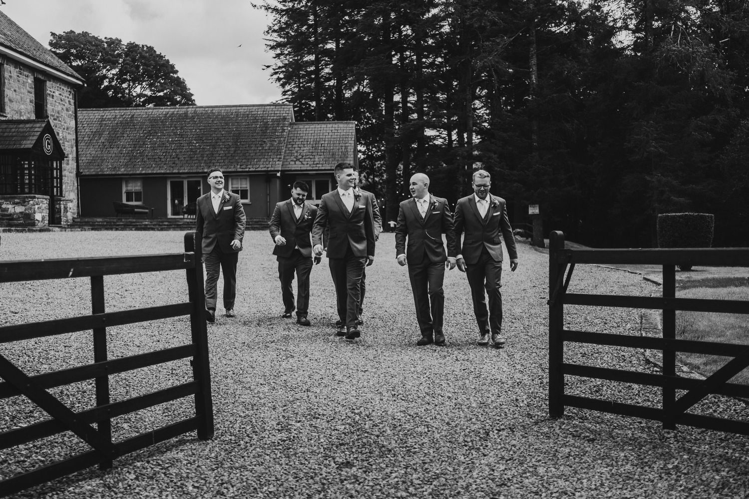 Groom and groomsmen walking together through open gates at a South Wales wedding venue, candid black and white moment before the ceremony.