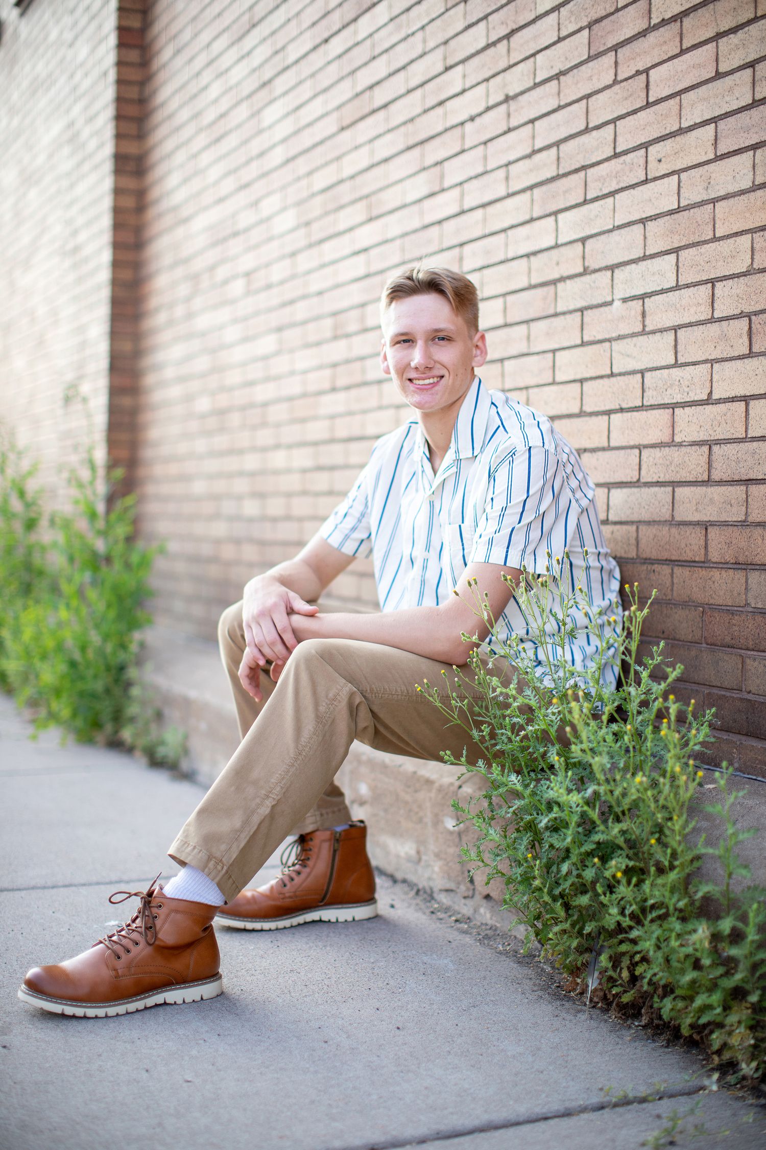 Person in striped shirt and khaki pants sits casually against brick wall with brown boots and small green shrubs nearby.