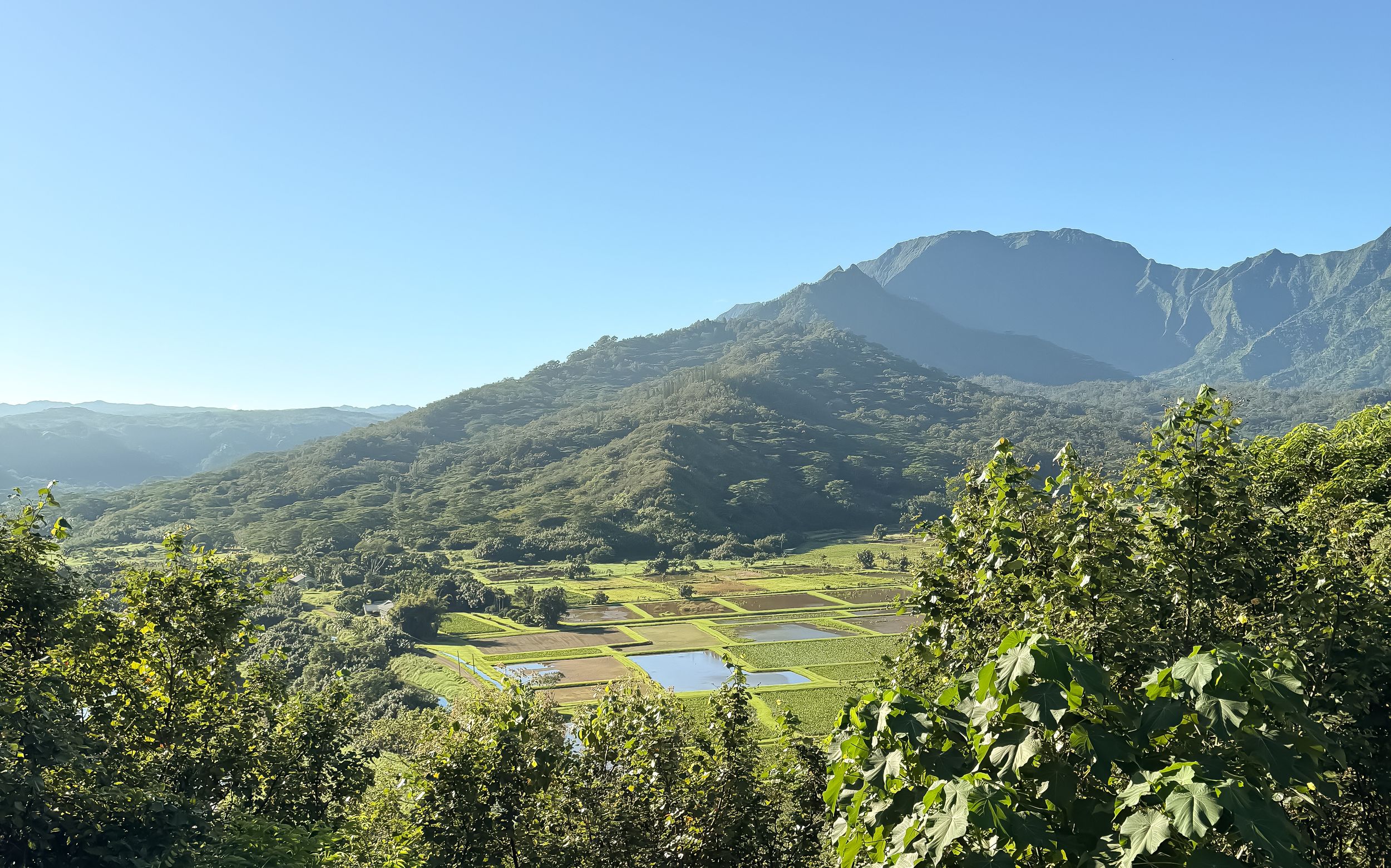 A panoramic mountain landscape with rolling green hills and a serene lake nestled among the peaks under blue skies.