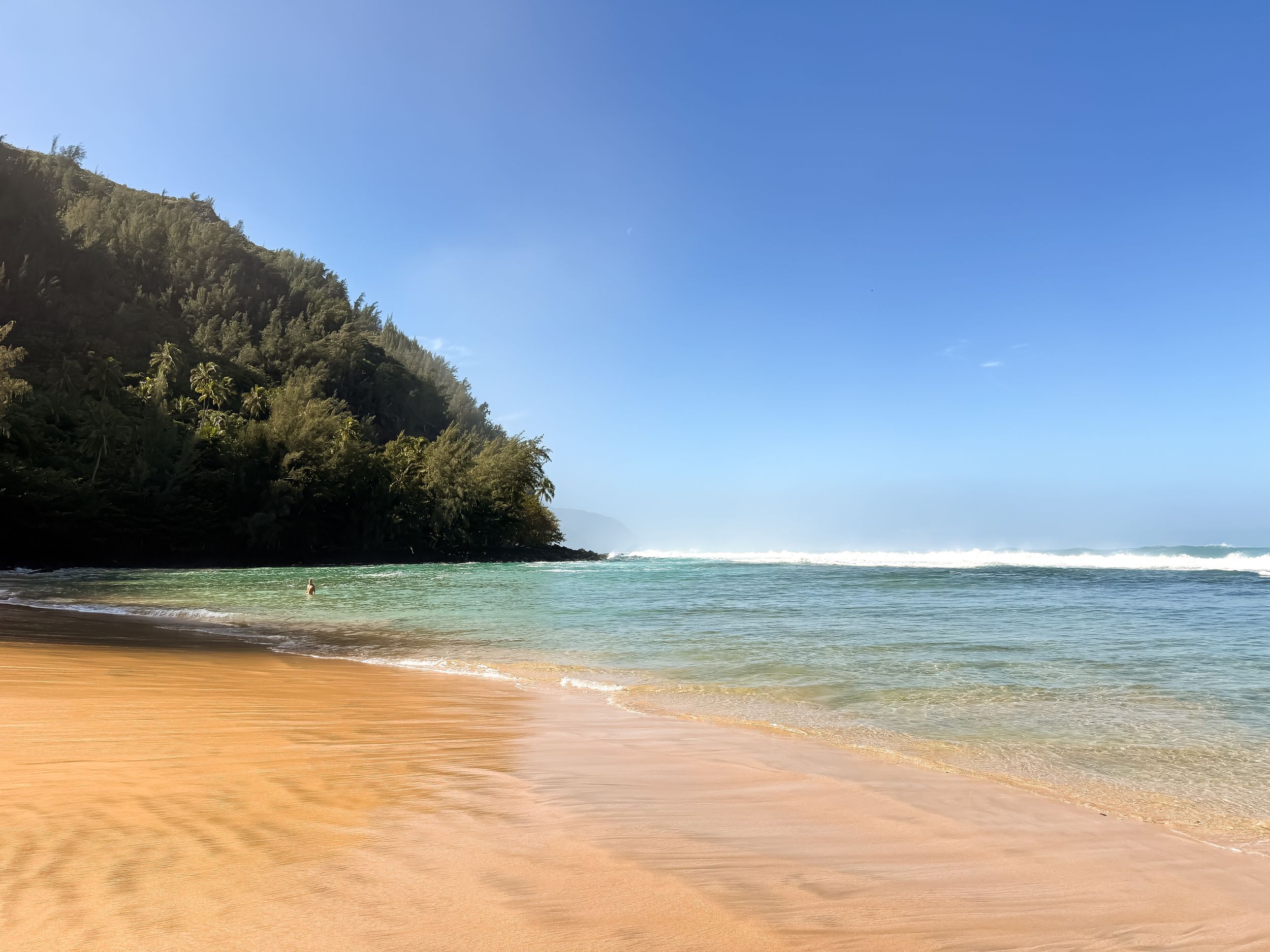 Turquoise ocean waves meet golden sand at a secluded beach cove with lush green cliffs in Hawaii.