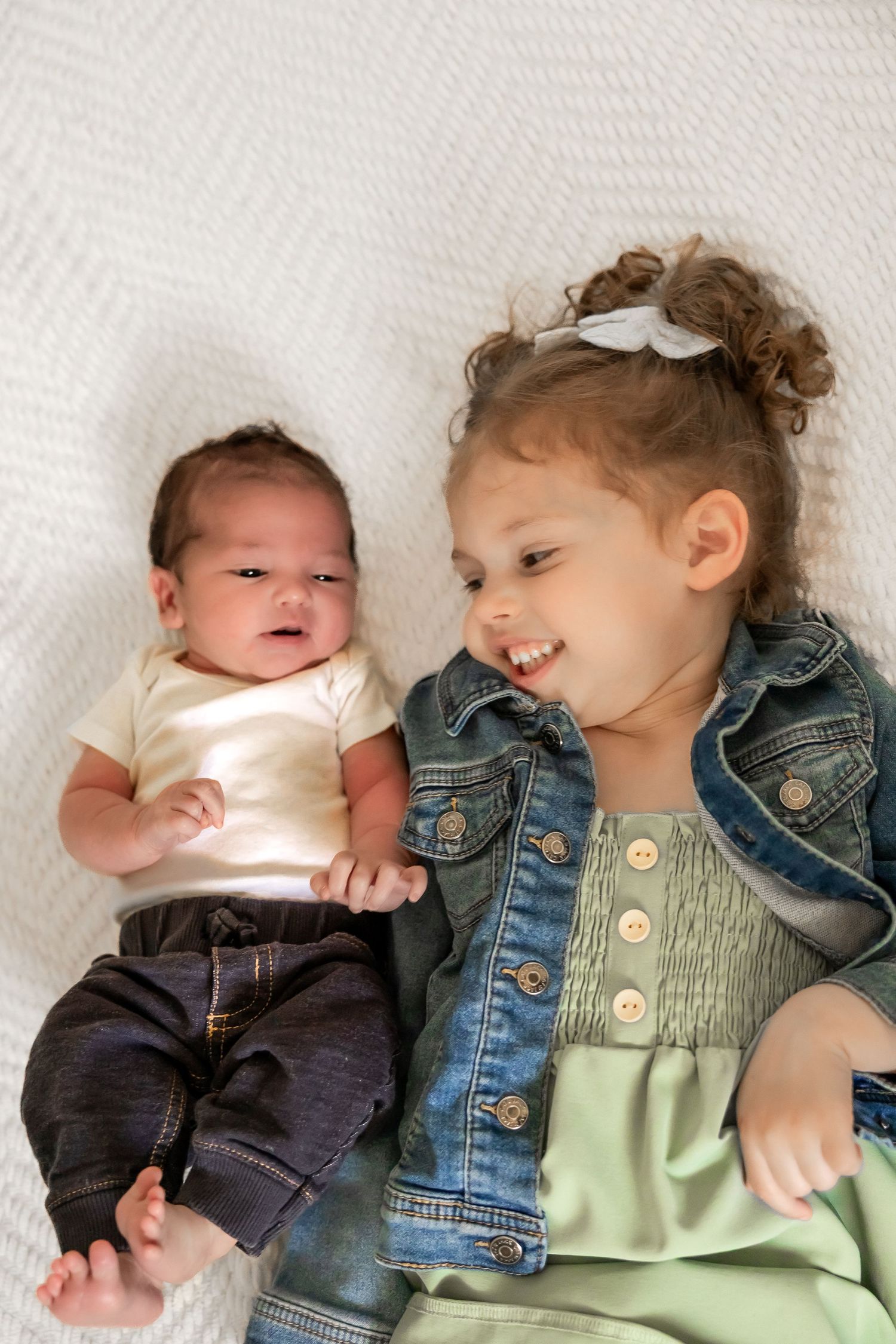 Two children laying on white fabric, with one wearing a denim jacket and mint dress and the other in casual attire.