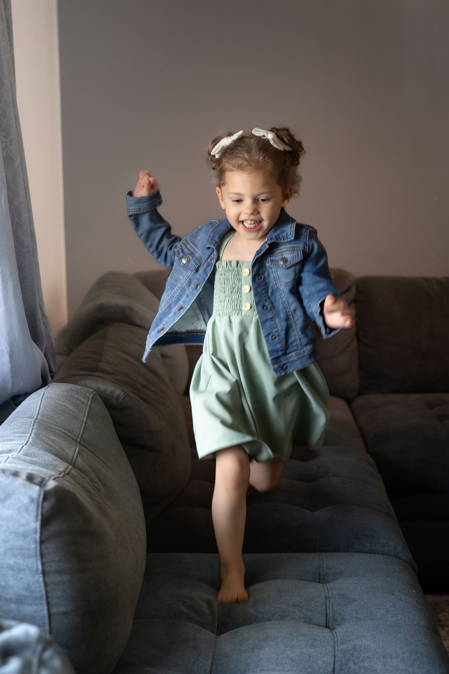 A sequence of four photos showing a toddler in a denim jacket jumping and playing on a dark gray couch.