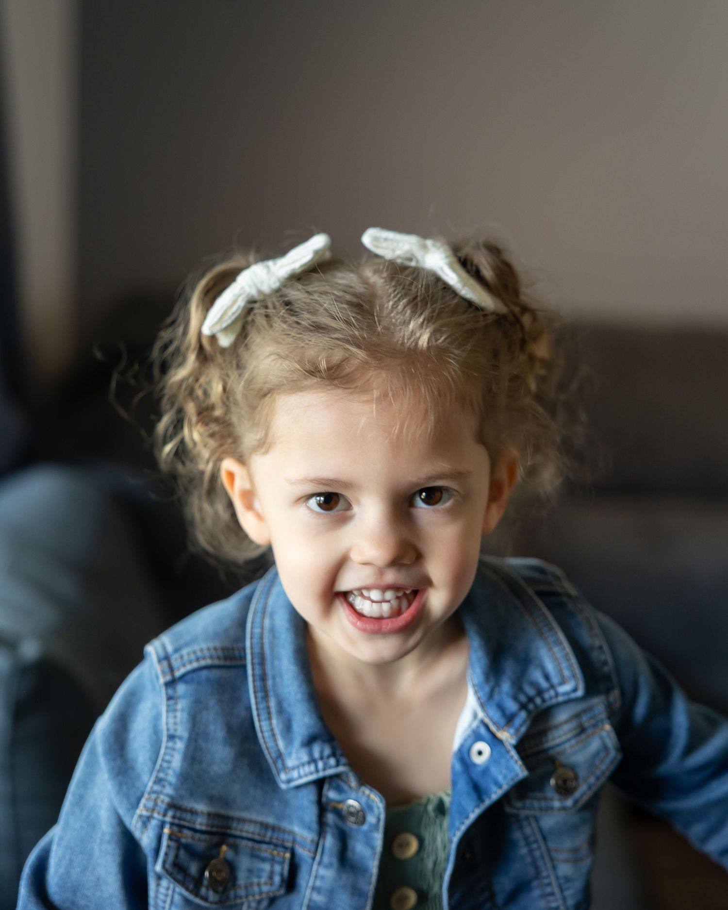 A joyful child wearing a denim jacket and white hair bows smiles brightly at the camera.
