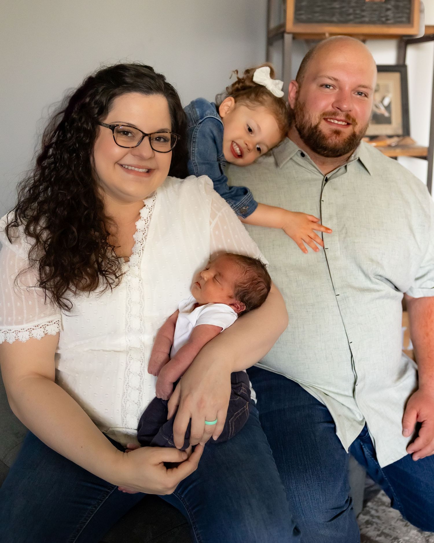 A family portrait showing parents with their newborn baby and toddler in a casual indoor setting.