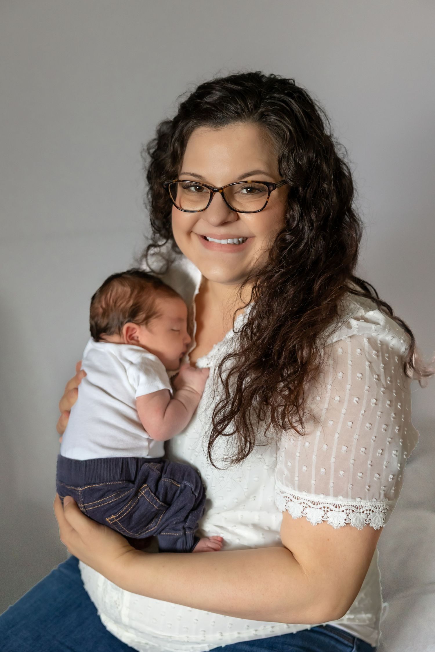 A tender moment captured between parent and baby in white lace dress against grey background.