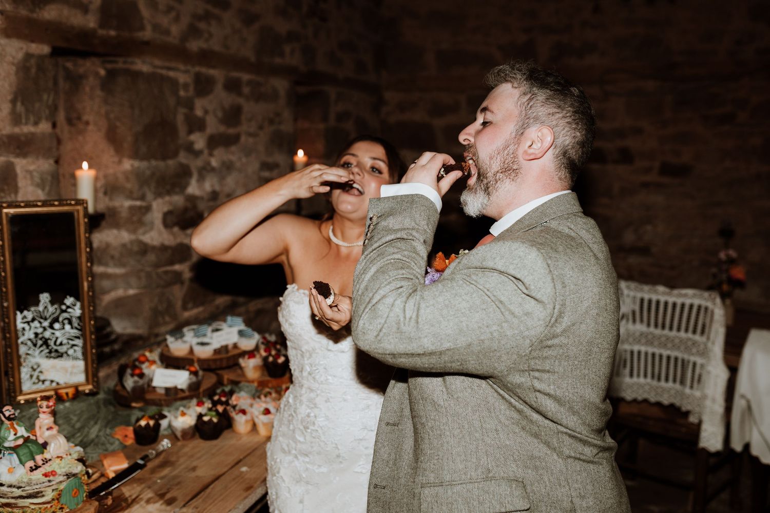 A couple eat cake at a rustic wedding reception with stone walls and candlelight.