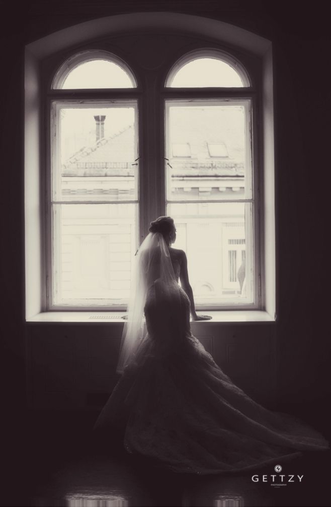 A silhouetted figure in a wedding dress gazes through an arched window in a moody black and white photo.