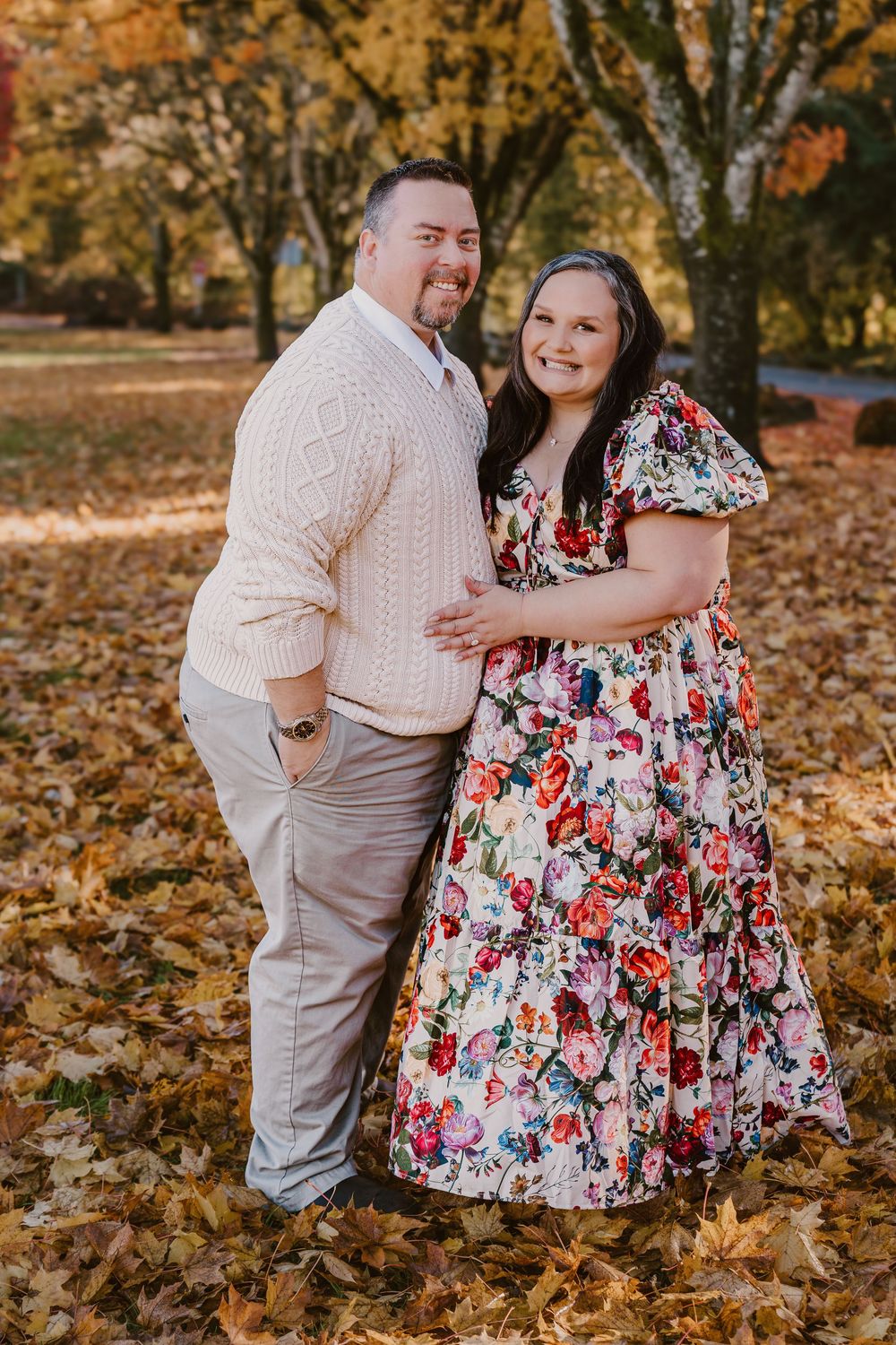 Couple posing together in autumn park, woman in floral dress, man in cream sweater, surrounded by fallen leaves.