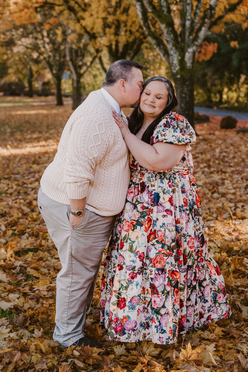 A couple shares a tender kiss among golden autumn leaves in a park, she wearing a floral dress, he in a cream sweater.