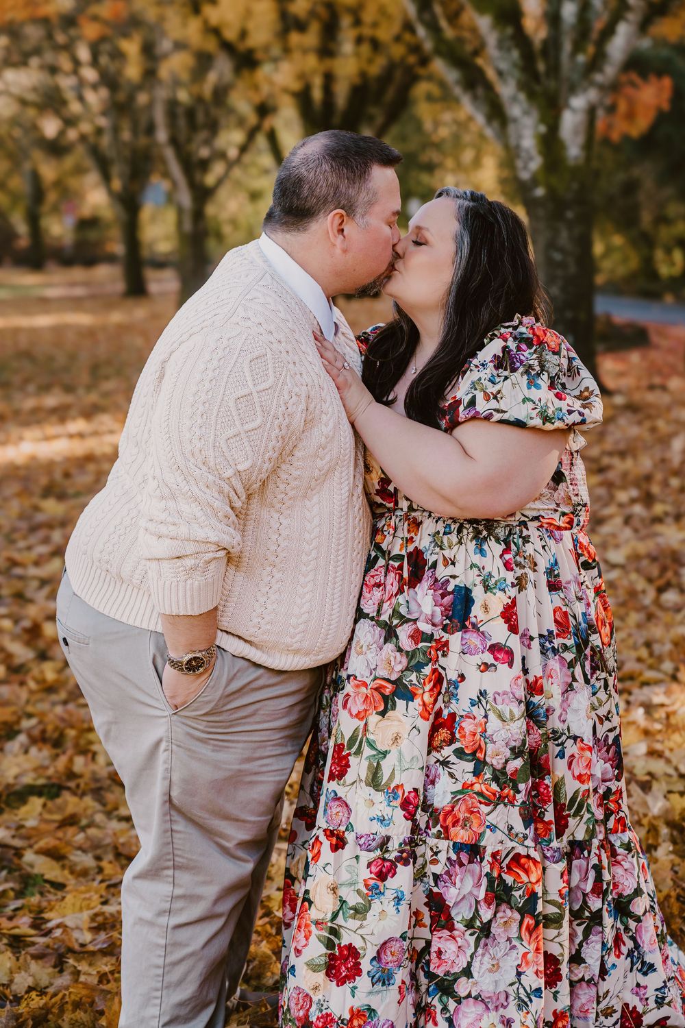 Couple sharing a romantic kiss in a park during autumn, wearing a floral dress and cream cable-knit sweater.