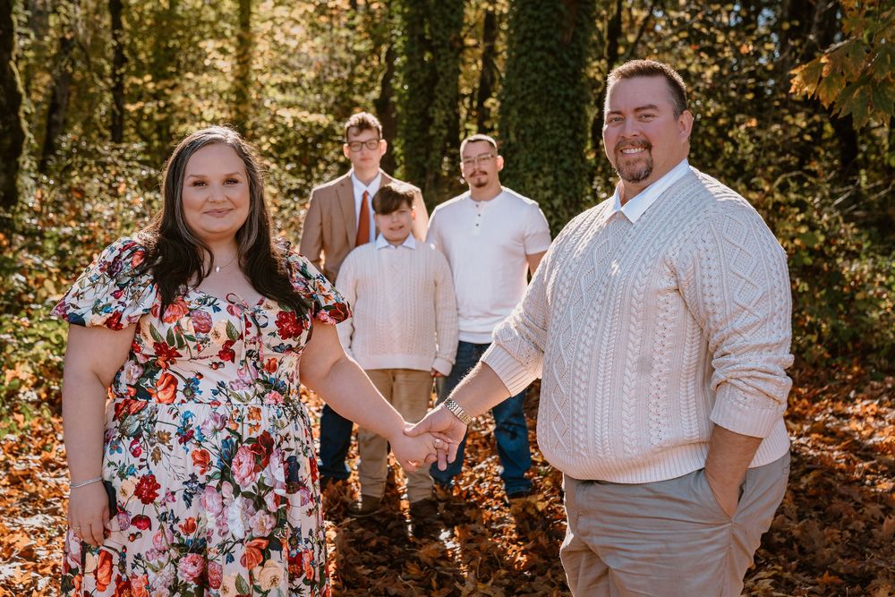 Family of five posing in autumn forest, parents holding hands in foreground, three children standing behind among colorful fall foliage.
