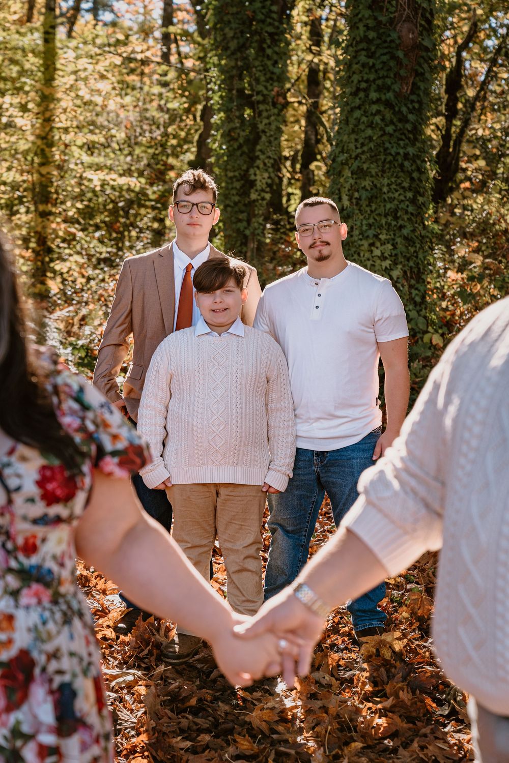 Family portrait in autumn forest with three males standing among fallen leaves, two adults holding hands in foreground.
