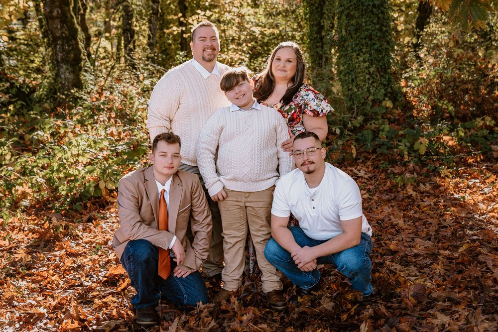 Family of five posing outdoors in autumn woods, surrounded by fallen leaves and lush green trees.