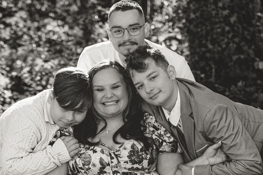 Black and white family portrait of four people smiling and embracing outdoors among green foliage.