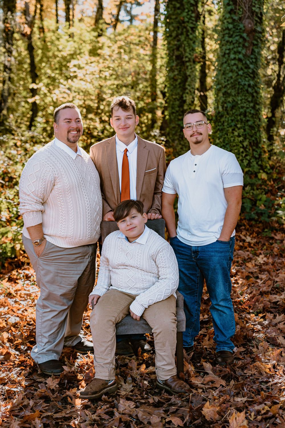 Four males pose for a family portrait outdoors among fallen autumn leaves with ivy-covered trees in the background.