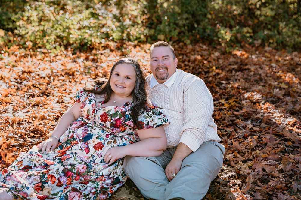 Couple smiling together outdoors, sitting on autumn leaves with colorful fall foliage in the background.