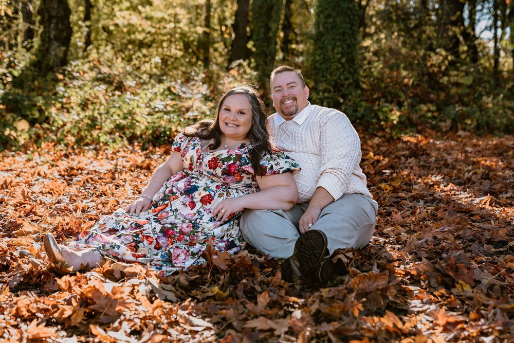 Couple sitting together in autumn leaves in a forest, woman in floral dress, man in white shirt, smiling.
