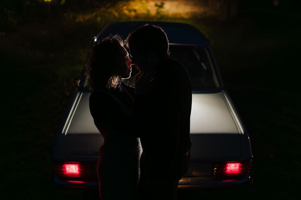 A silhouette of a couple stands in front of car headlights creating a romantic backlit scene at night.