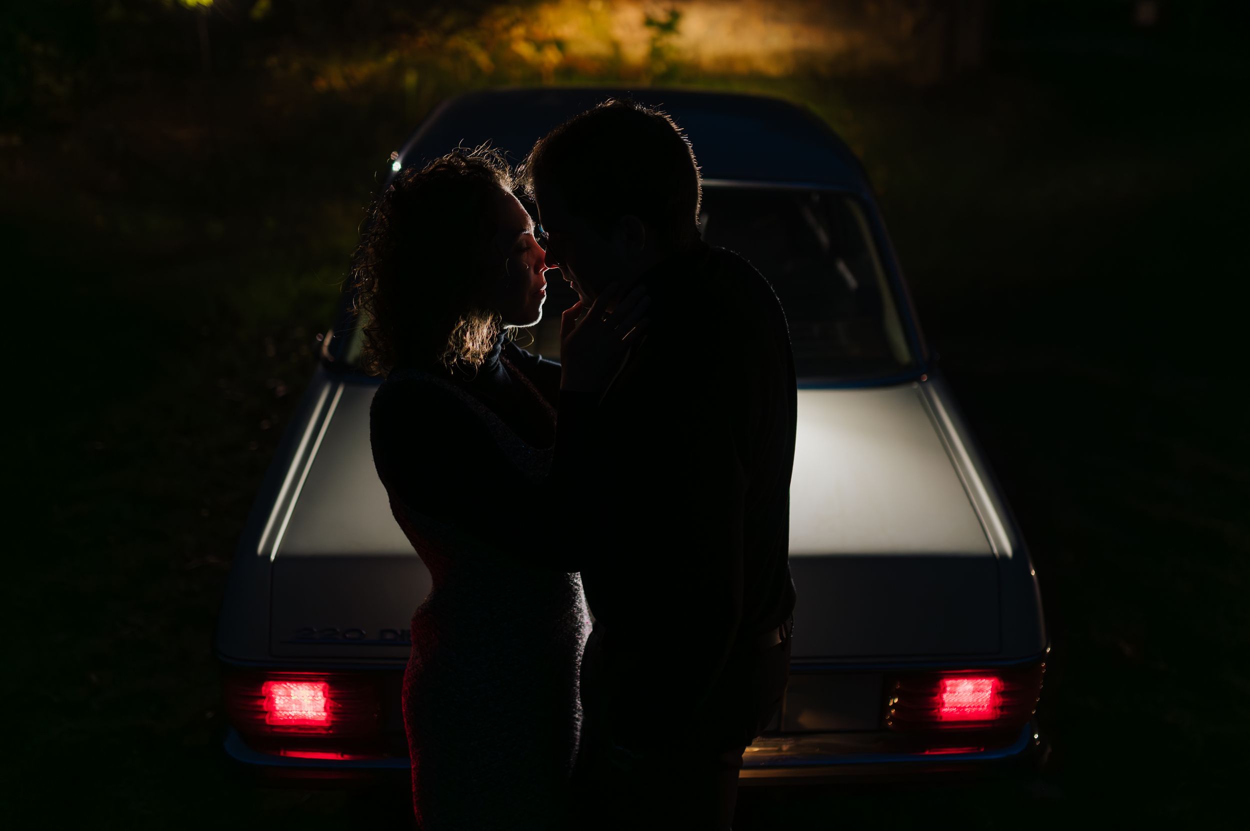A silhouette of a couple stands in front of car headlights creating a romantic backlit scene at night.