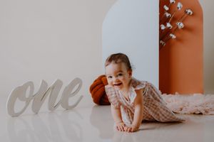 Baby in white dress lying on floor with decorative 'one' text in background for first birthday photoshoot.