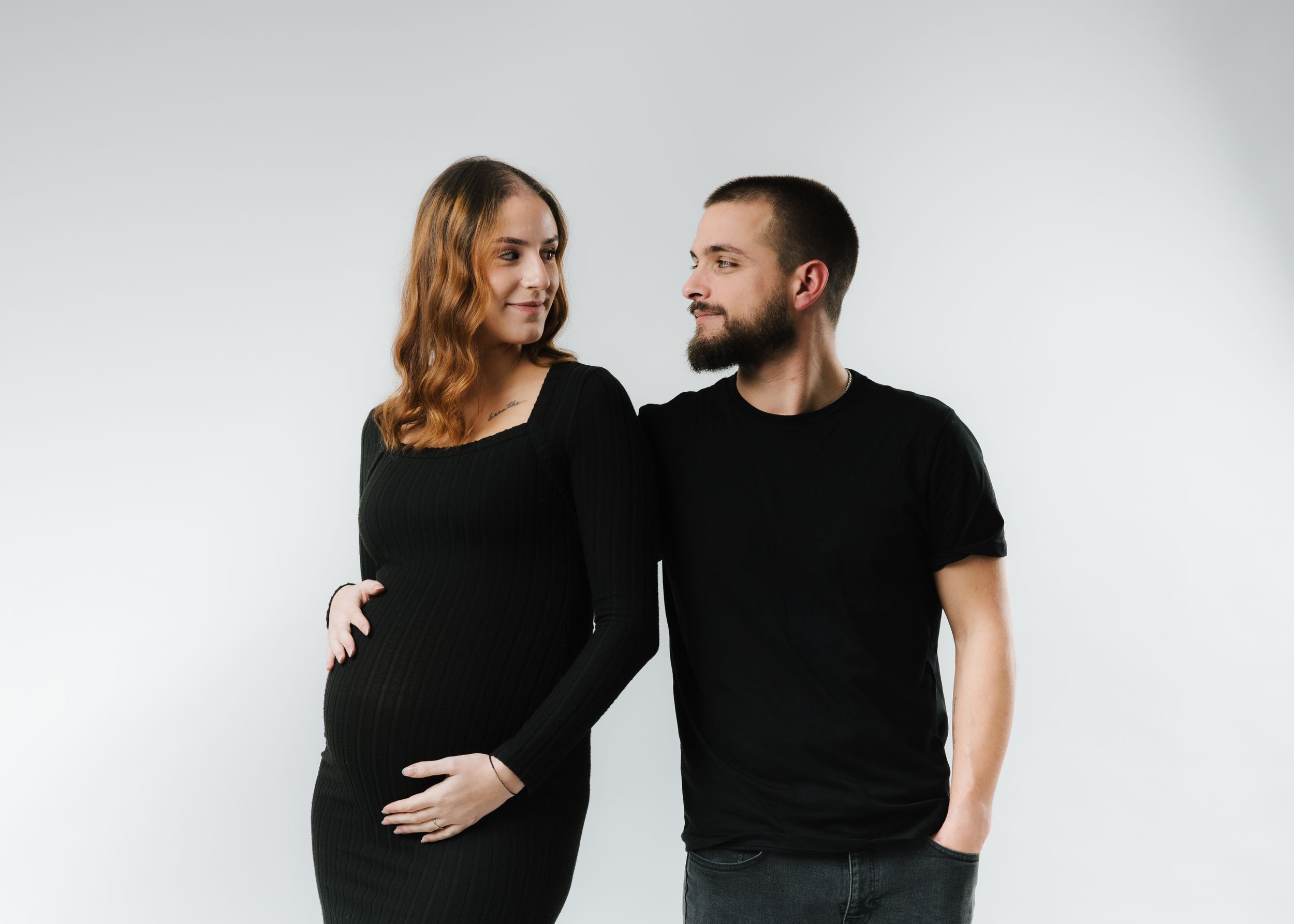 A couple wearing black outfits poses together in a minimalist studio setting for a maternity photoshoot.