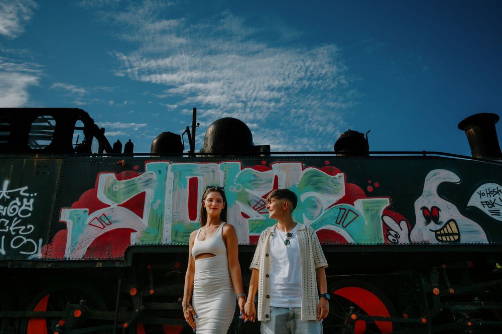 A group of people stand in front of a colorful mural wall with graffiti art and red accents against a blue sky.