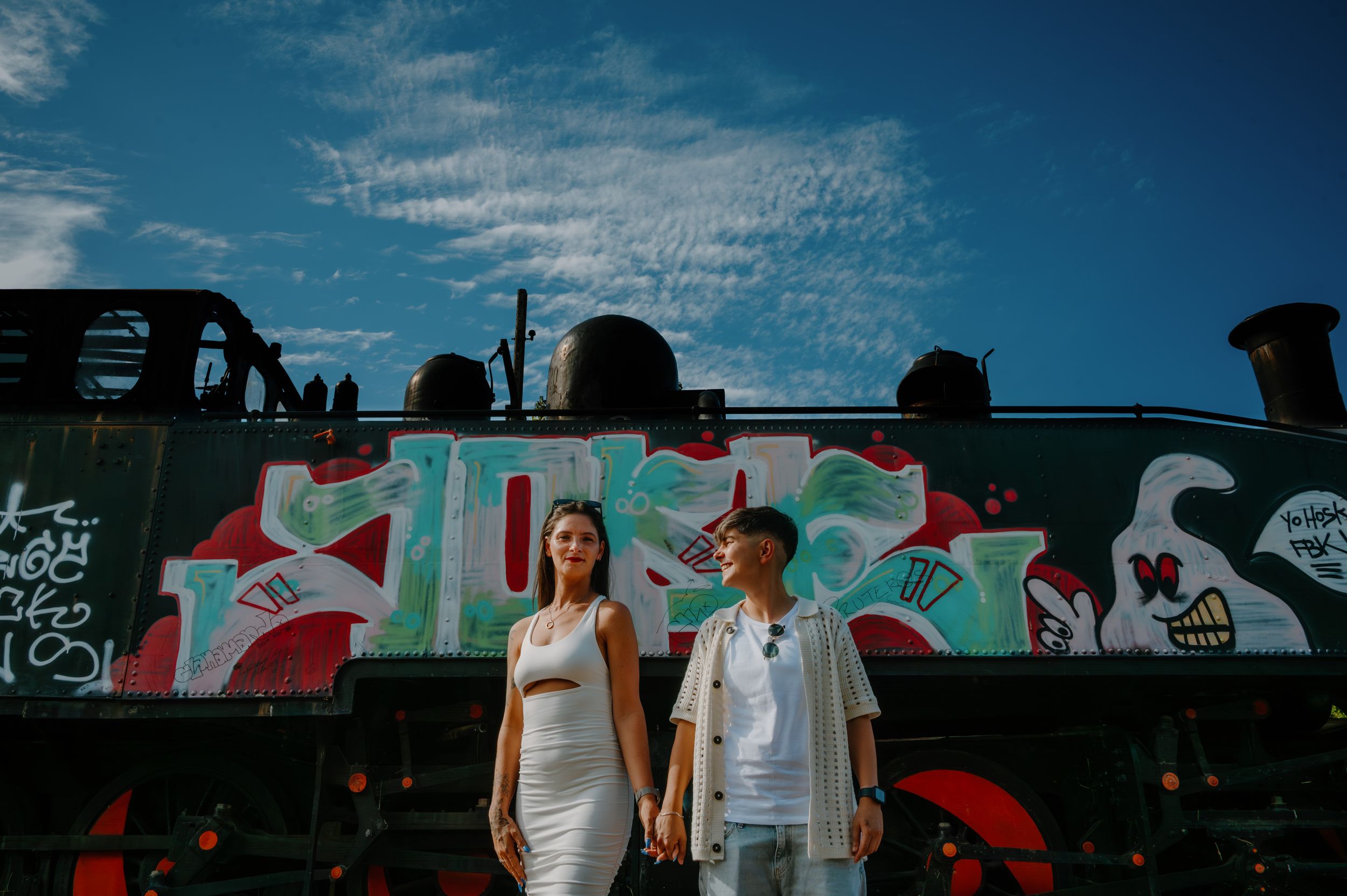 A group of people stand in front of a colorful mural wall with graffiti art and red accents against a blue sky.