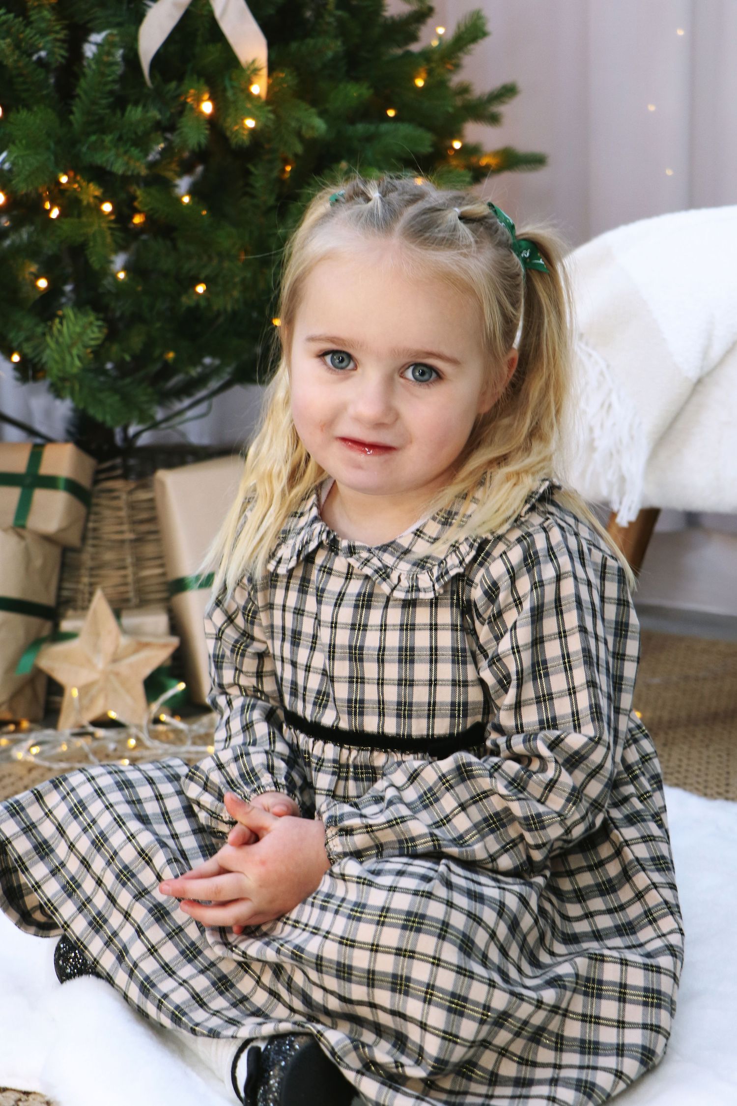 A young child in a black and white plaid dress sits by a decorated Christmas tree with twinkling lights.